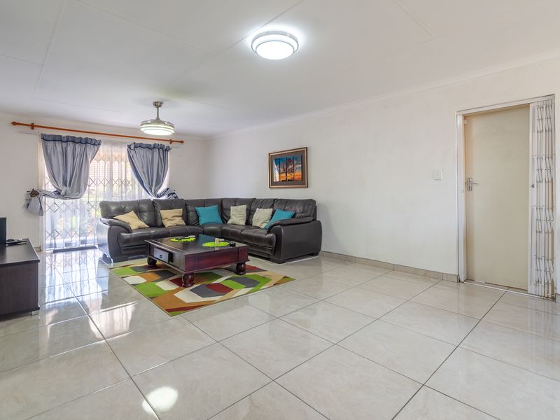 A spacious living room featuring neutral tiled flooring, a ceiling fan, and a sliding door with security gate access to the exterior.