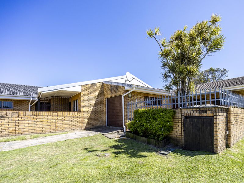 Front exterior view of a single-story brick house featuring a garage, boundary wall with security fencing, and a paved driveway.