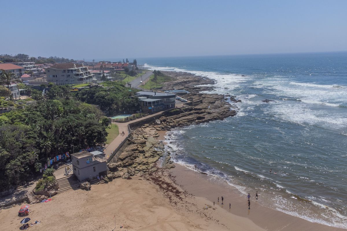 An aerial view of the coastal property location, showcasing the beach, rocky shoreline, and surrounding residential apartment buildings.