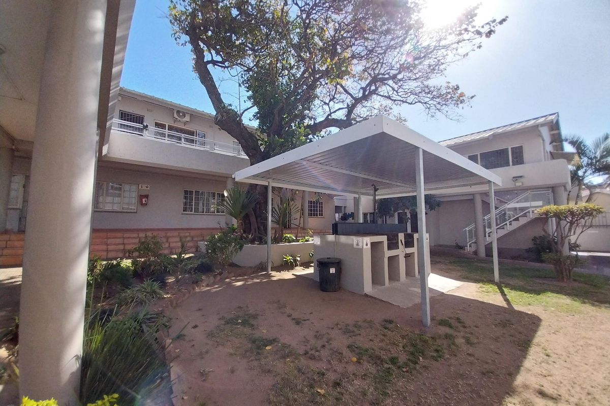 Communal outdoor area featuring a covered built-in braai station, paved ground, and established garden beds within the apartment complex.