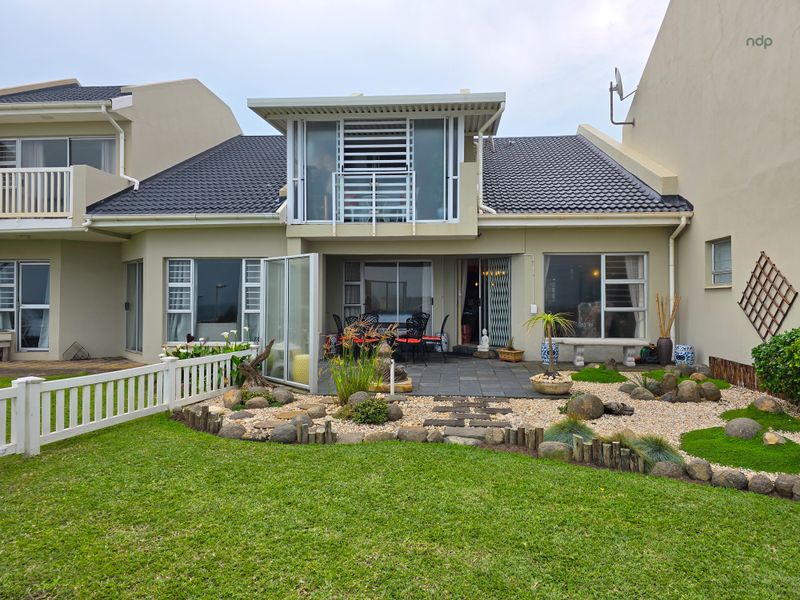 Rear view of the property featuring a landscaped garden, stone pathway, tiled patio area, and an upper-level balcony with sliding glass doors.