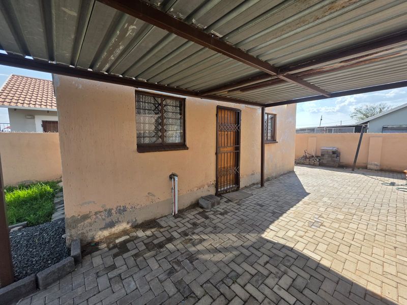 Rear exterior view featuring a paved area, covered carport structure, and security-fitted windows and door, showing signs of wear on the exterior wall finish.