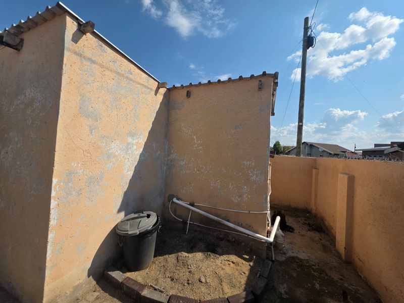 Rear exterior area showing weathered walls, exposed plumbing pipes, and a boundary wall.