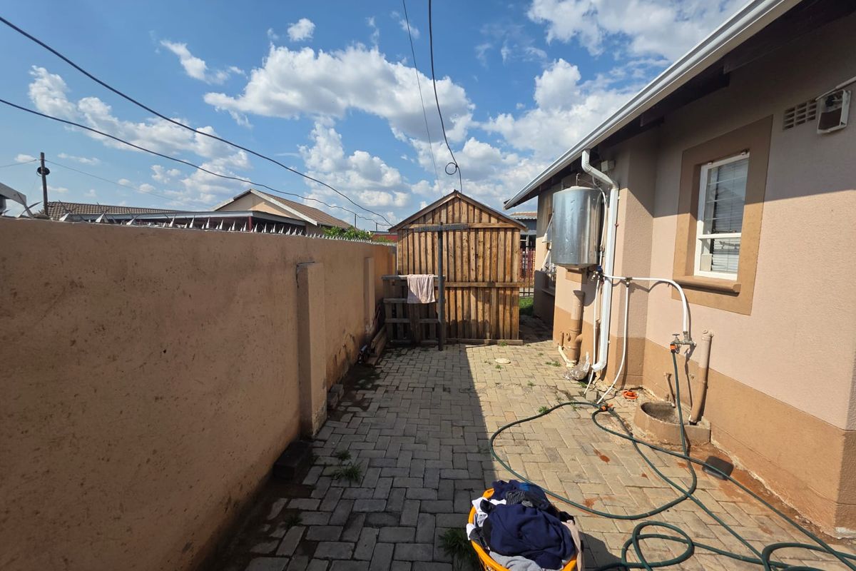 A functional rear courtyard featuring paved flooring, a boundary wall with electric fencing, an external geyser, and a wooden storage shed.