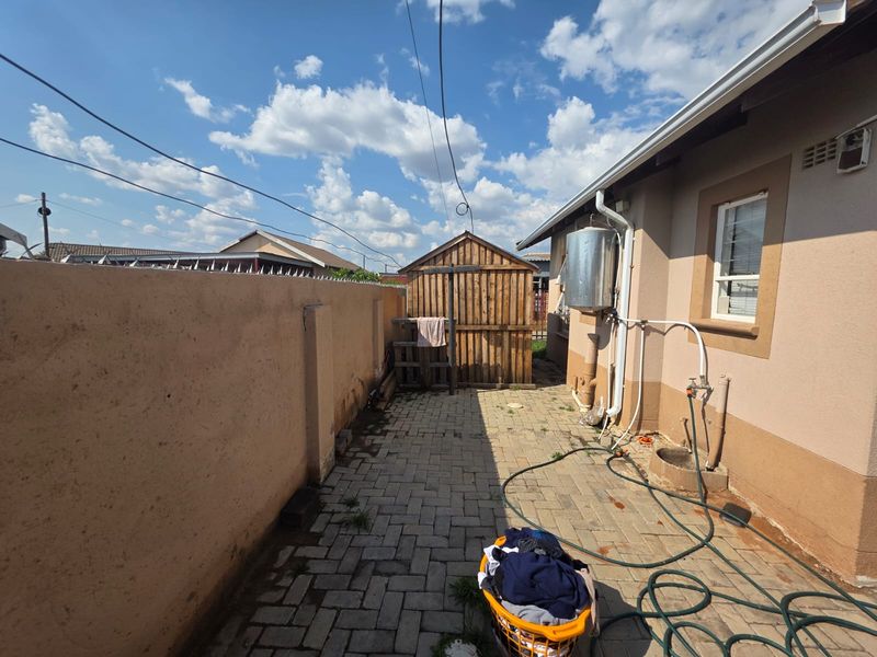 A functional rear courtyard featuring paved flooring, a boundary wall with electric fencing, an external geyser, and a wooden storage shed.