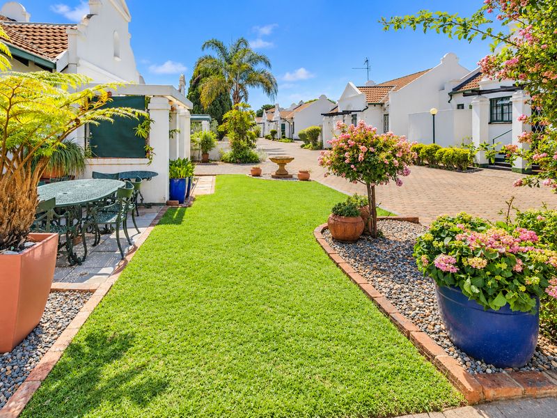 A well-maintained retirement village exterior featuring a manicured lawn, paved driveway, stone garden borders, and a charming covered patio area.