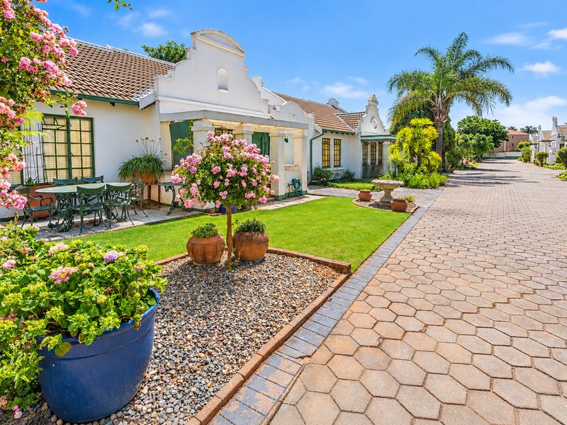 A well-maintained retirement village exterior featuring Cape Dutch architectural elements, a paved driveway, and a manicured garden with a covered patio area.