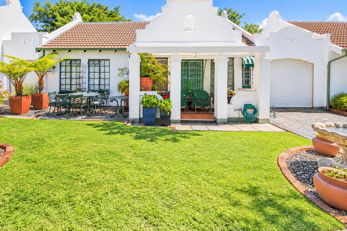A well-maintained rear exterior view featuring a covered patio, manicured lawn, stone fountain, and attached garage with a classic Cape Dutch architectural style.