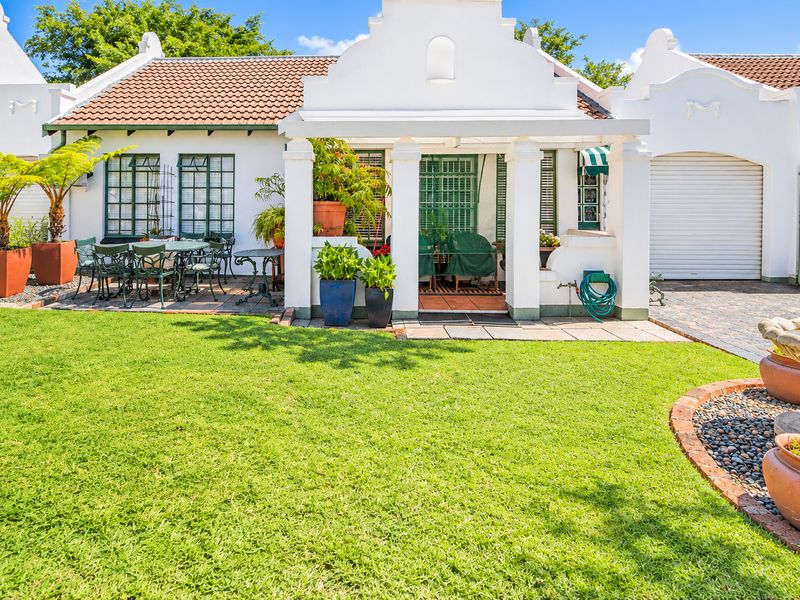 A well-maintained rear exterior view featuring a covered patio, manicured lawn, stone fountain, and attached garage with a classic Cape Dutch architectural style.