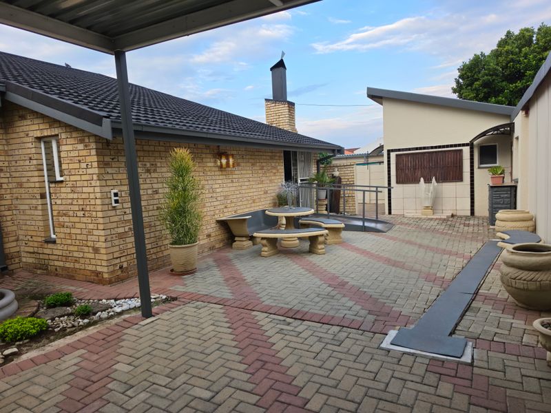 A well-maintained paved patio area featuring a brick facade, chimney, and a metal carport structure with an accessible wheelchair ramp.