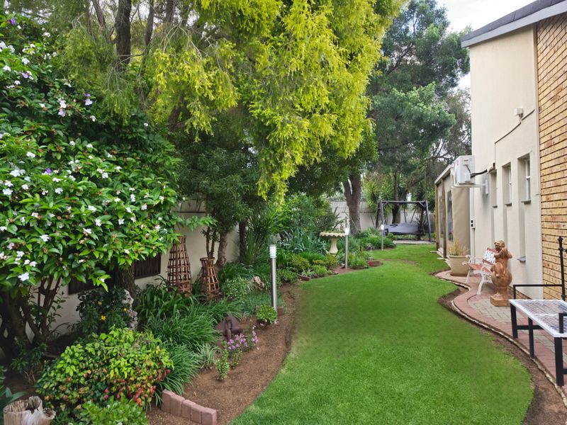 A well-maintained rear garden featuring a lush green lawn, established trees, landscaped garden beds with brick edging, and a paved side walkway.