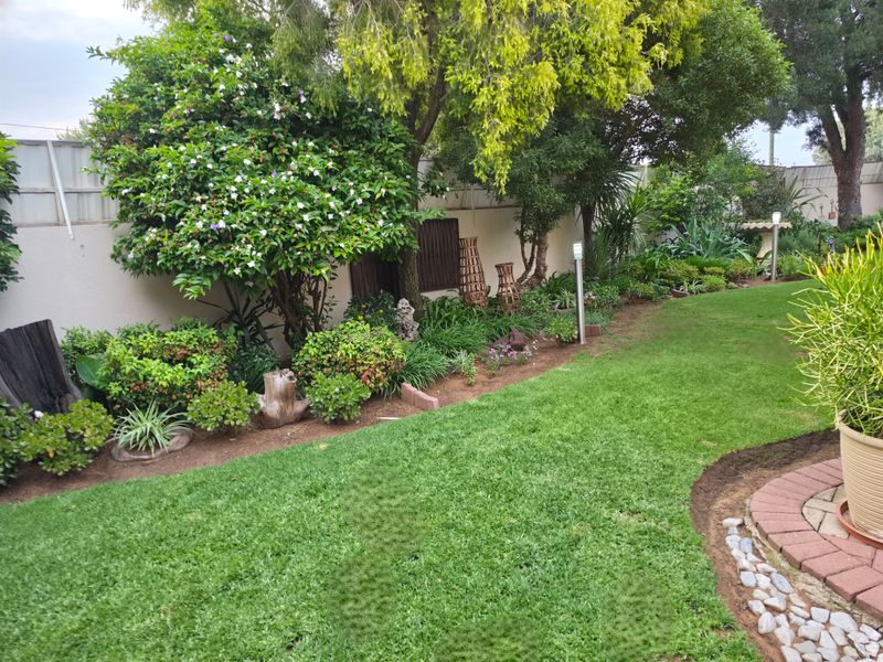 A well-maintained residential garden featuring a lush green lawn, mature trees, established shrubbery, and neat brick-edged garden beds.