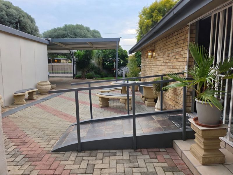 A paved outdoor patio area featuring a metal accessibility ramp with handrails, leading to a sliding door entrance alongside a face brick wall.