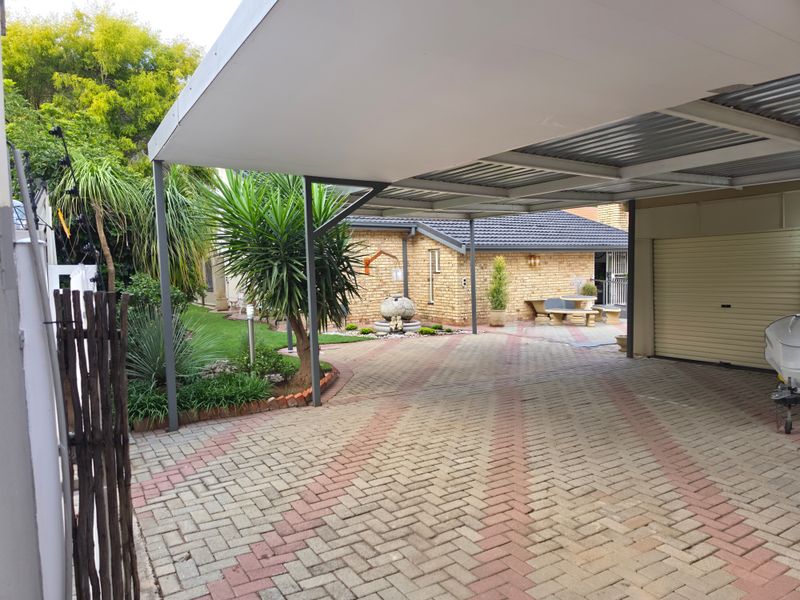 A well-maintained front exterior featuring a spacious paved driveway, a covered carport, and a neat landscaped garden with brick facade detailing.