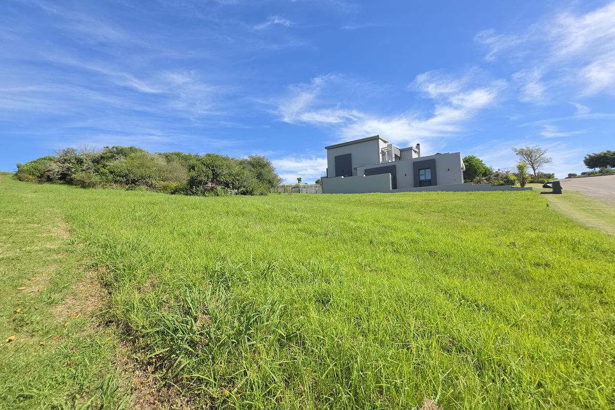 A view of a vacant residential plot with a gentle slope, featuring a modern house in the background and established greenery.