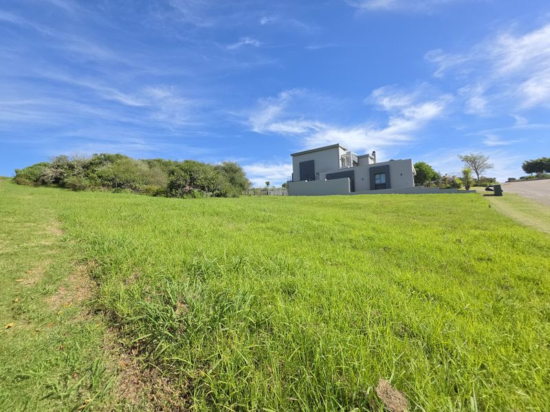 A view of a vacant residential plot with a gentle slope, featuring a modern house in the background and established greenery.
