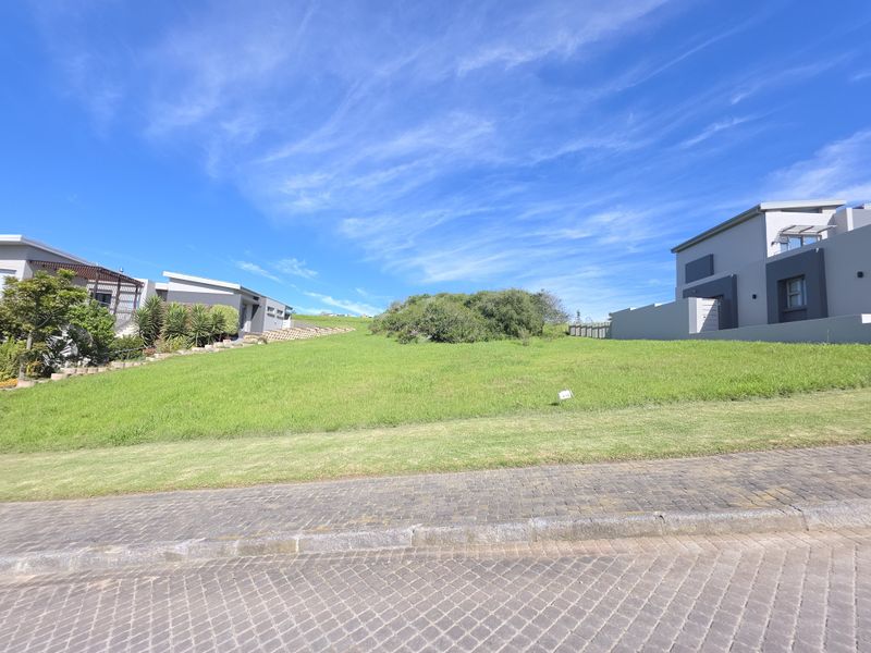 A vacant residential plot featuring a well-maintained grass lawn, situated between existing modern homes with paved street access.