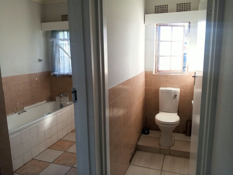 A dated bathroom featuring a built-in bathtub and separate toilet area with original ceramic tiling and natural light through window openings.