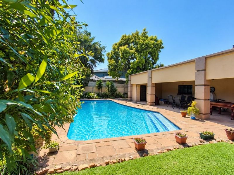 A well-maintained outdoor entertainment area featuring a swimming pool, paved deck, and a covered patio with a built-in braai.