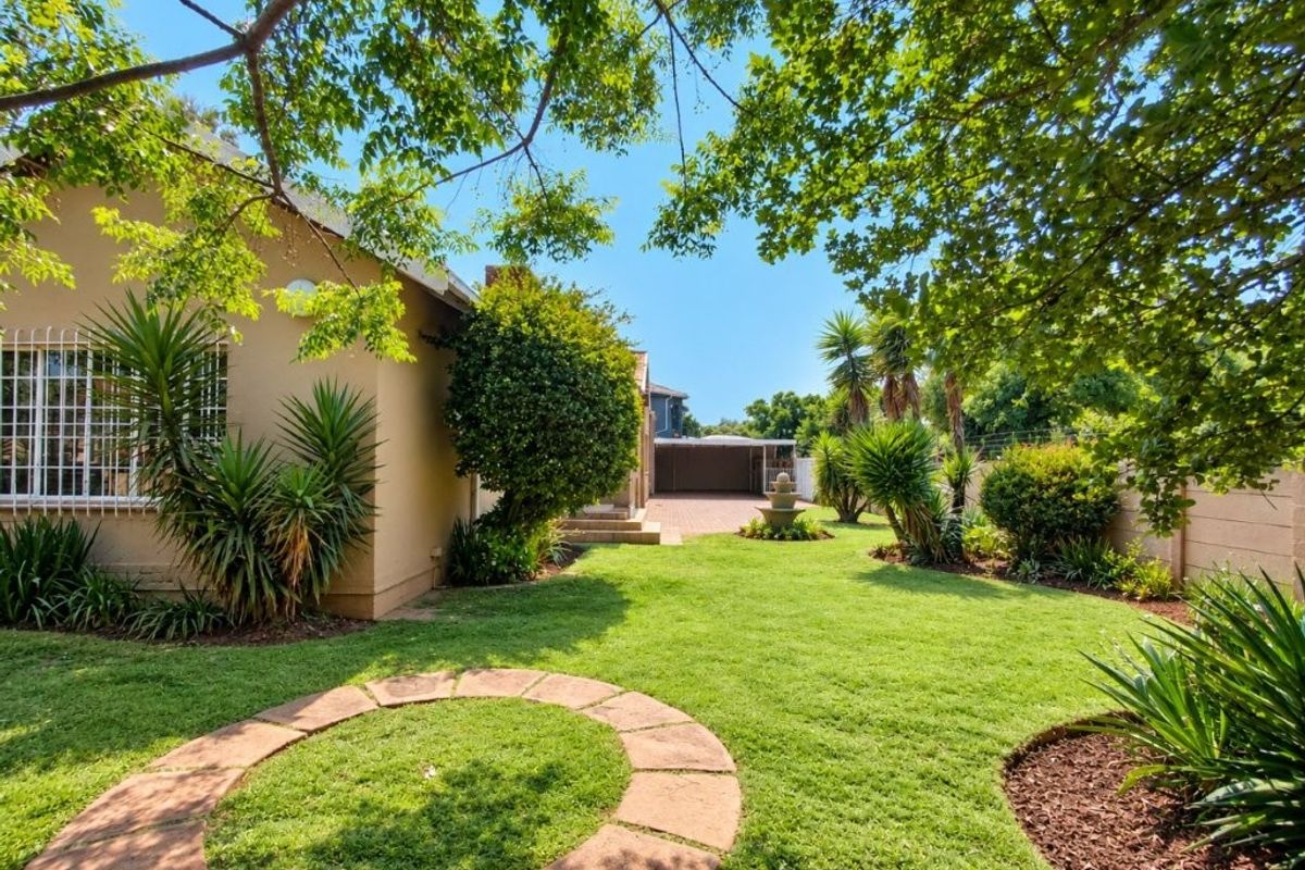A well-maintained rear garden featuring a lush lawn, mature trees, stone fountain, and brick-paved driveway leading to a garage.