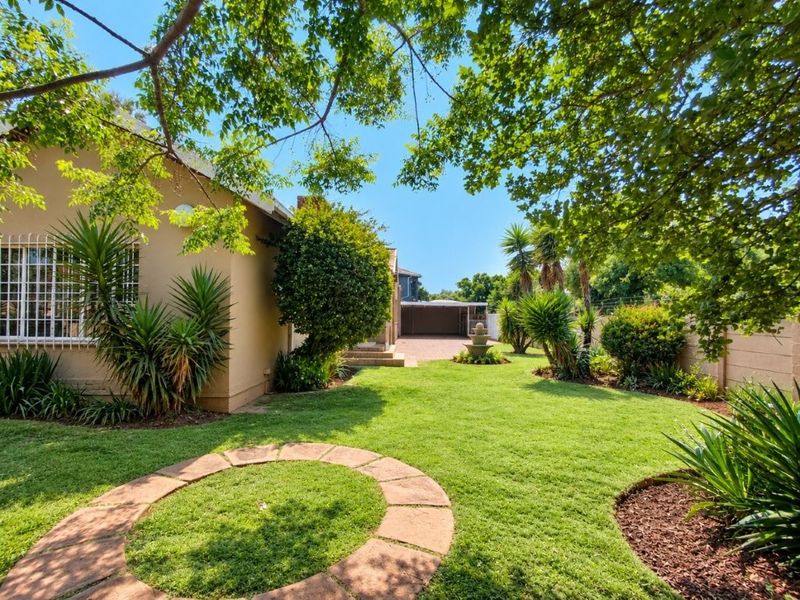 A well-maintained rear garden featuring a lush lawn, mature trees, stone fountain, and brick-paved driveway leading to a garage.