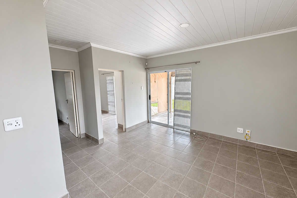 A bright, empty living area featuring neutral tiled floors, a white tongue and groove ceiling, and a sliding door leading to an outdoor space.