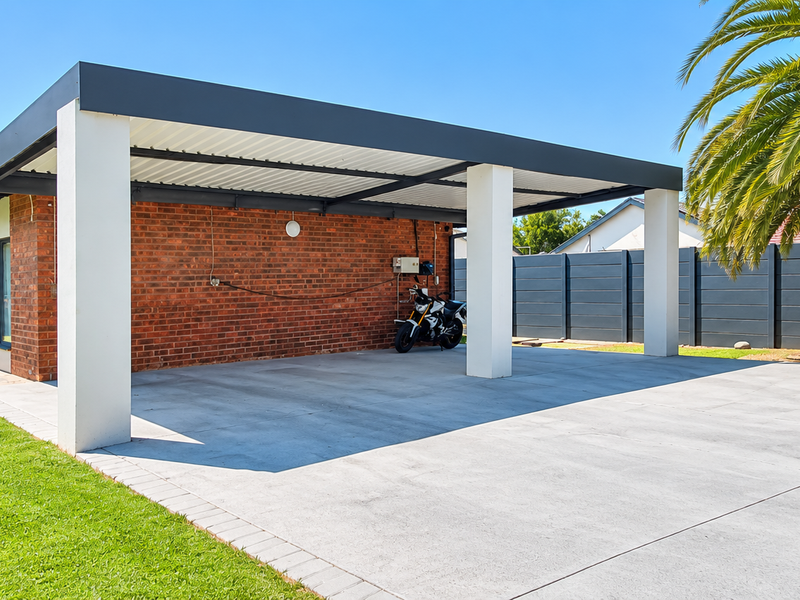 A spacious, modern carport with a concrete driveway, brick exterior, and a built-in outdoor fireplace, set within a well-maintained garden area.