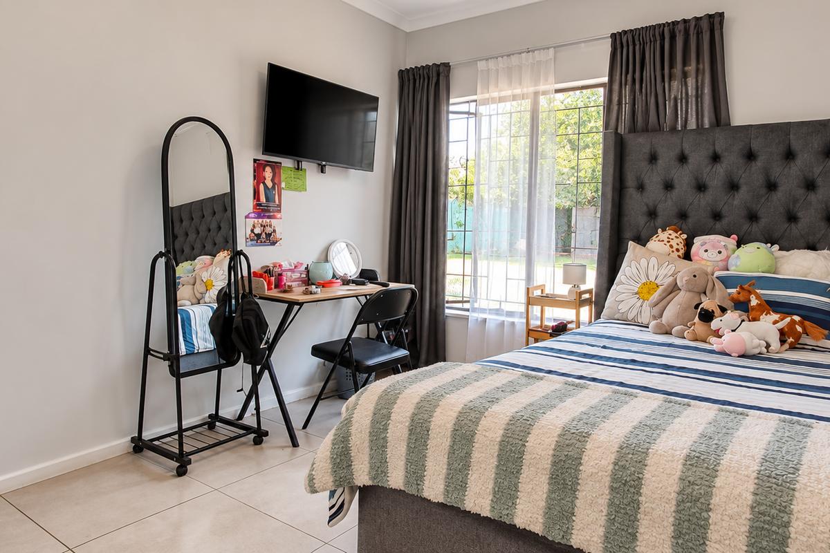 A well-maintained bedroom featuring neutral wall tones, tiled flooring, and a large window with security bars, providing natural light and a view of the garden.