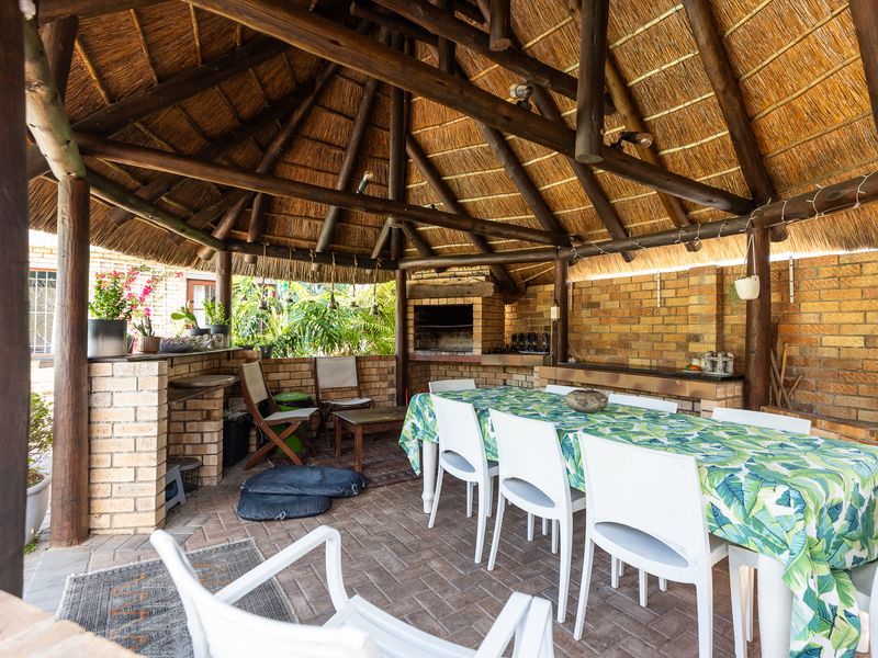 Spacious covered outdoor patio featuring a traditional thatched roof, exposed timber beams, and a built-in brick braai, perfect for alfresco dining and entertaining.