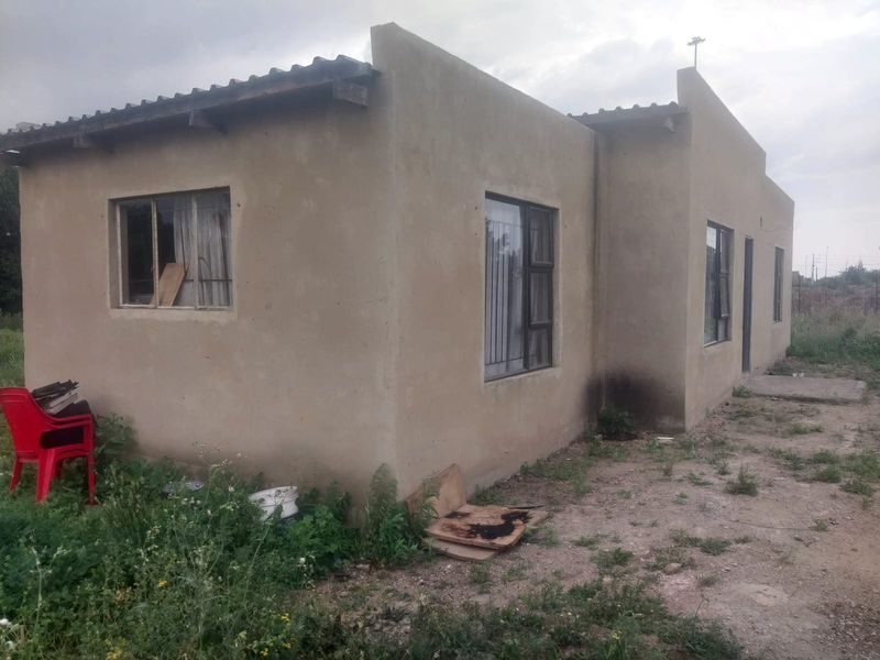 Rear view of a single-story residential structure featuring plastered masonry walls, metal window frames, and corrugated roof sheeting, set on an unlandscaped plot.