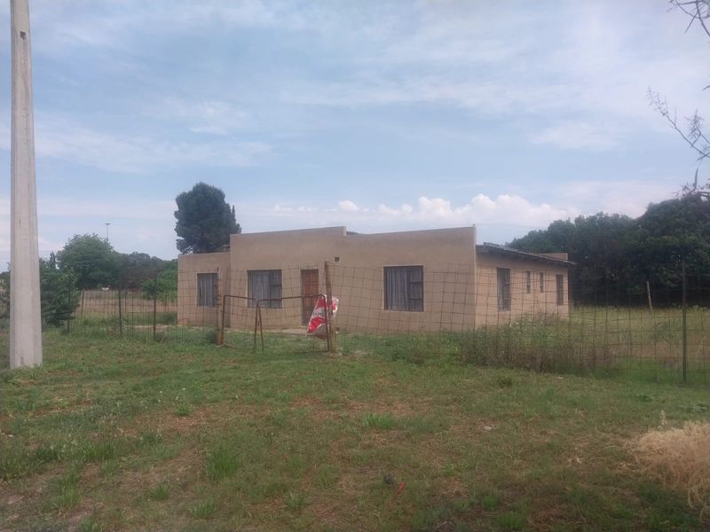 A single-story residential property featuring a flat roof and wire mesh perimeter fencing, set on a large, unmaintained plot of land.