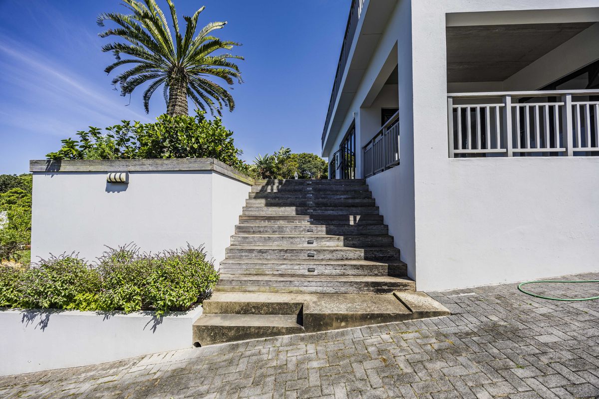 Rear exterior view featuring a timber staircase leading to an elevated balcony, set against a landscaped garden with a paved driveway.