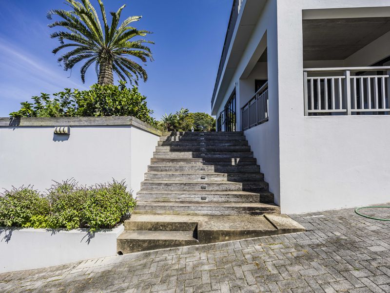 Rear exterior view featuring a timber staircase leading to an elevated balcony, set against a landscaped garden with a paved driveway.