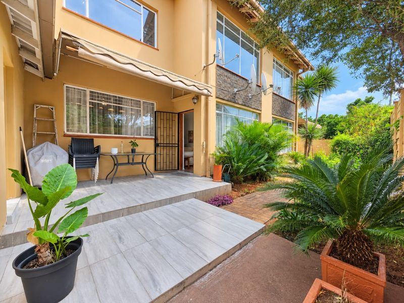 Rear view of the apartment featuring a tiled patio area with a retractable awning, security gate access, and landscaped garden surroundings.