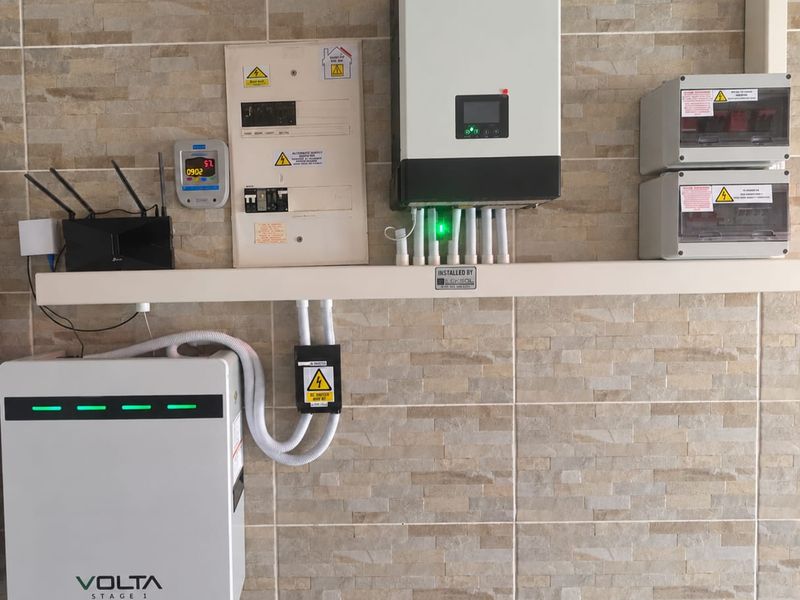 A clean utility area featuring a modern solar inverter, battery storage system, and electrical distribution board mounted on a stone-tiled wall.