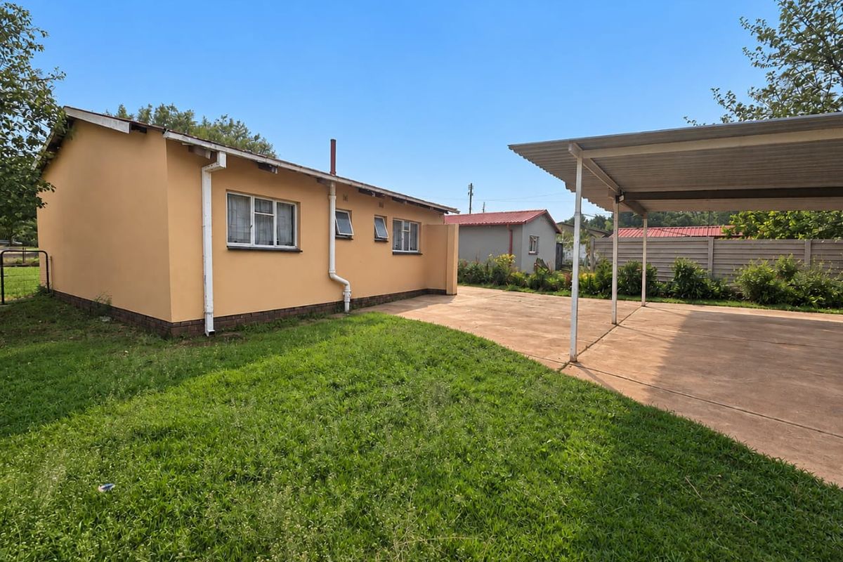 Rear view of the property featuring a paved driveway, a metal carport, and a lawn area enclosed by a precast concrete boundary wall.