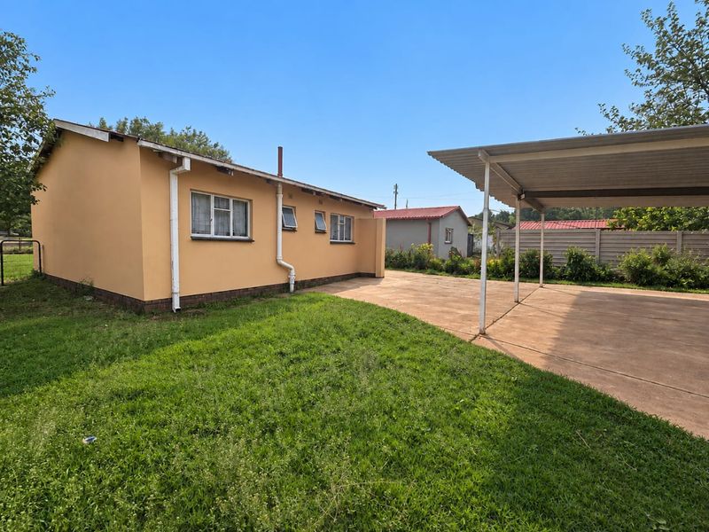 Rear view of the property featuring a paved driveway, a metal carport, and a lawn area enclosed by a precast concrete boundary wall.