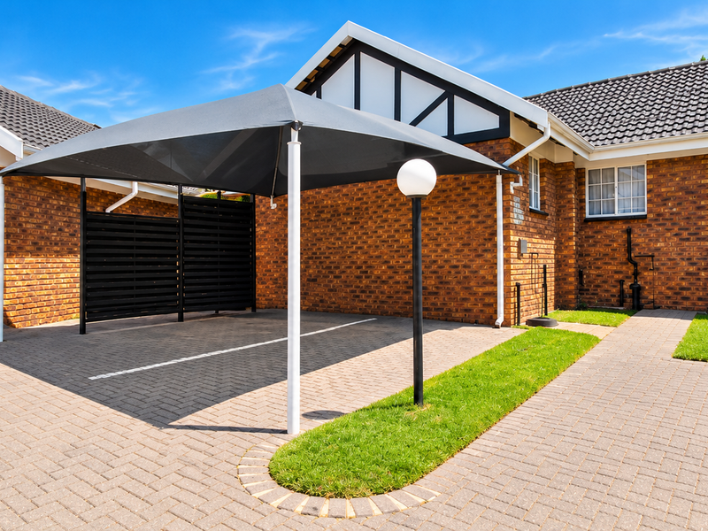 Front exterior view of a townhouse featuring a brick facade, tiled roof, paved driveway, and a covered parking area with a shade sail.