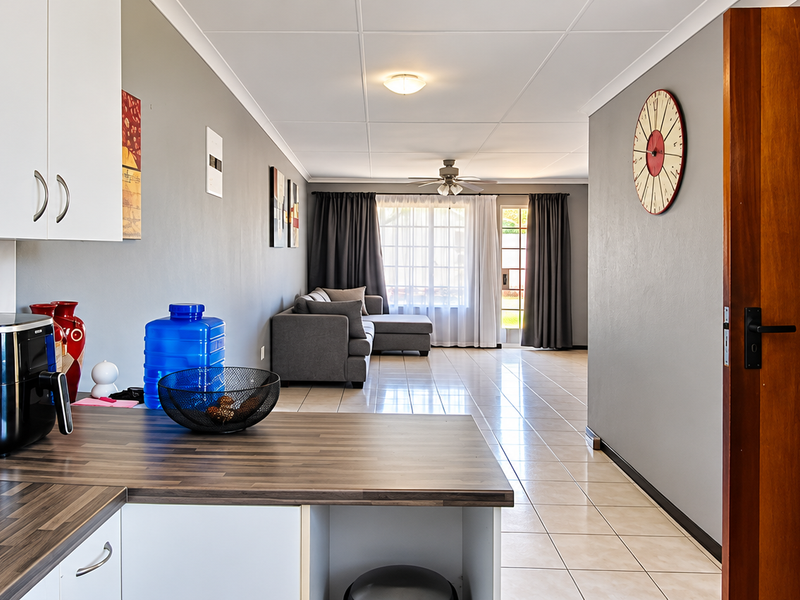 An open-plan living and kitchen area featuring tiled flooring, white cabinetry with laminate countertops, and a ceiling fan, leading to a bright outdoor access point.