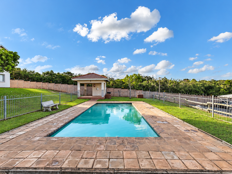 A well-maintained communal swimming pool area featuring a paved deck, a covered gazebo, and secure perimeter fencing within a landscaped estate setting.