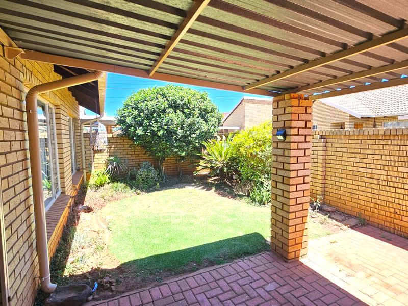 A covered patio area featuring brick paving, a sturdy brick pillar, and a corrugated metal roof, overlooking a private garden with facebrick boundary walls.