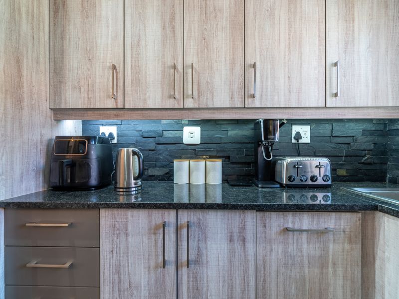 Modern kitchen featuring light wood-grain cabinetry, dark granite countertops, and a textured stone backsplash with integrated under-cabinet lighting.