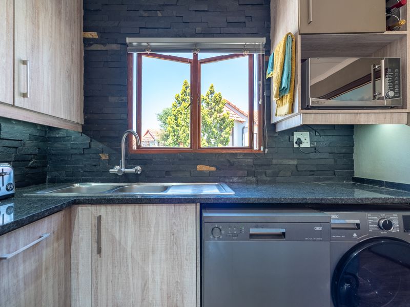 A modern kitchen area featuring light wood-grain cabinetry, dark stone countertops, a slate tile backsplash, and a functional stainless steel sink and tap.