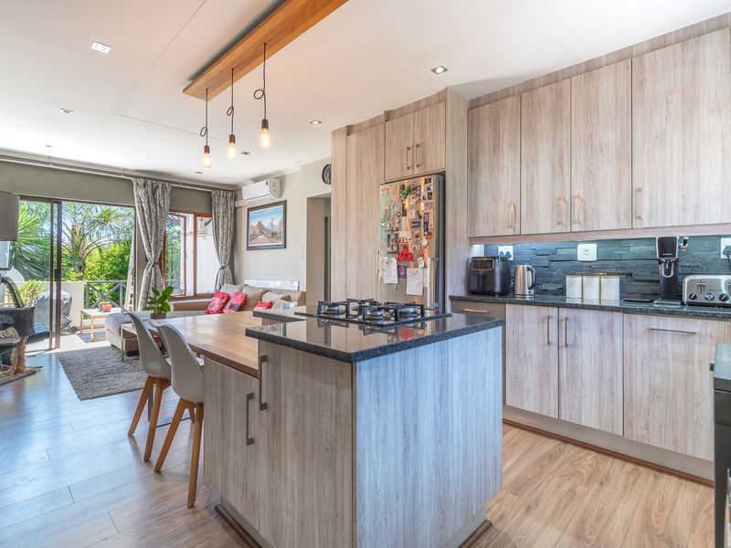 Modern open-plan kitchen and living area featuring a central island with gas hob, light wood cabinetry, stone splashback, and seamless flow to a balcony.