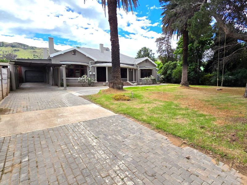 Front view of a single-story house featuring a paved driveway, covered carport, attached garage, and a spacious front garden with mature trees.