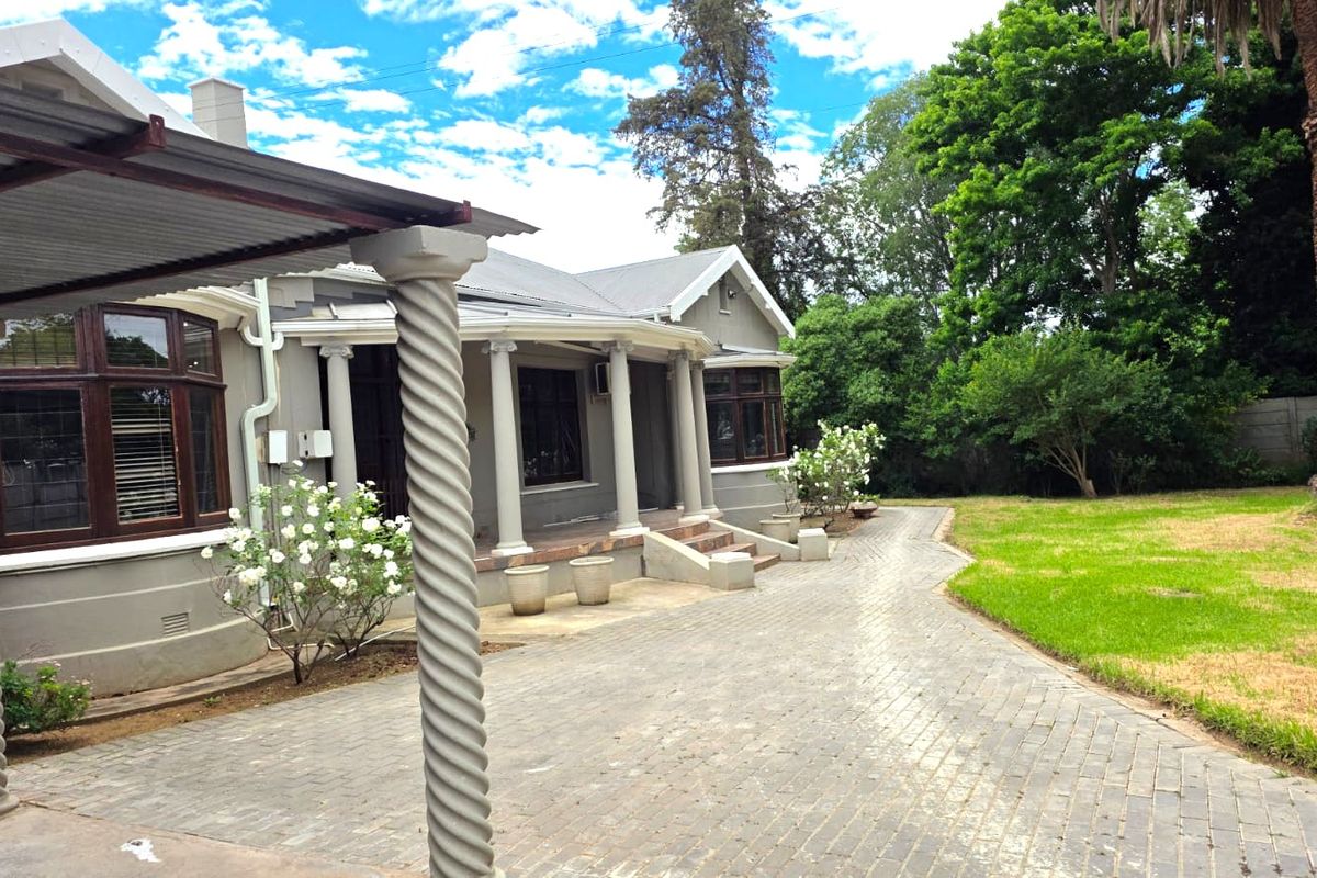 A rear view of the house featuring a covered patio with decorative columns, paved driveway, and established garden surroundings.