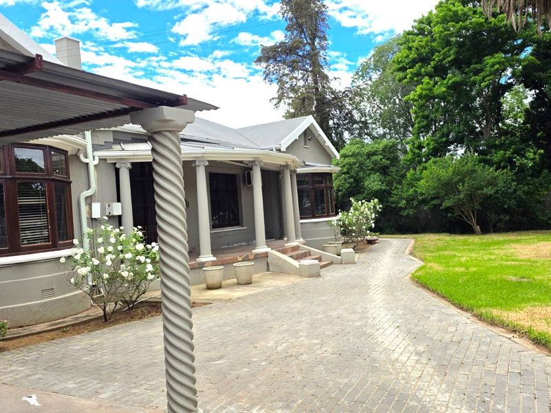 A rear view of the house featuring a covered patio with decorative columns, paved driveway, and established garden surroundings.