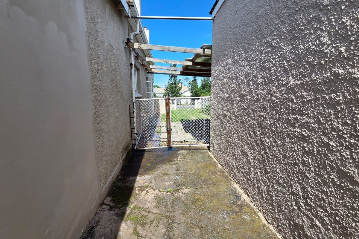 A narrow side passage featuring a concrete walkway, a chain-link gate, and a weathered timber pergola structure leading to the rear garden area.