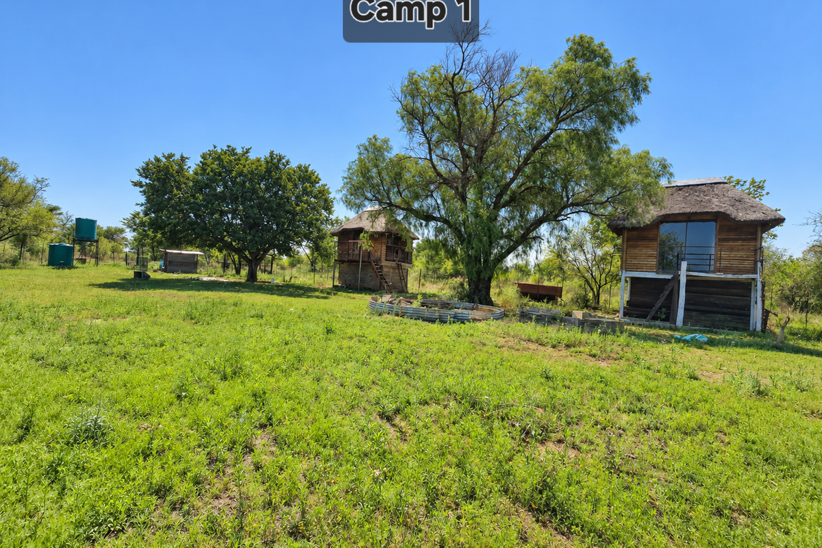A rural farm camp area featuring elevated thatched-roof cabins, water storage tanks, and open grazing land under a clear sky.