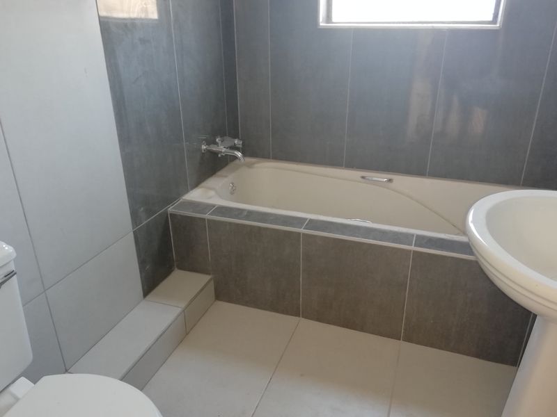 A functional bathroom featuring a built-in bathtub with chrome fixtures, a pedestal basin, and a toilet, finished with neutral wall and floor tiling.